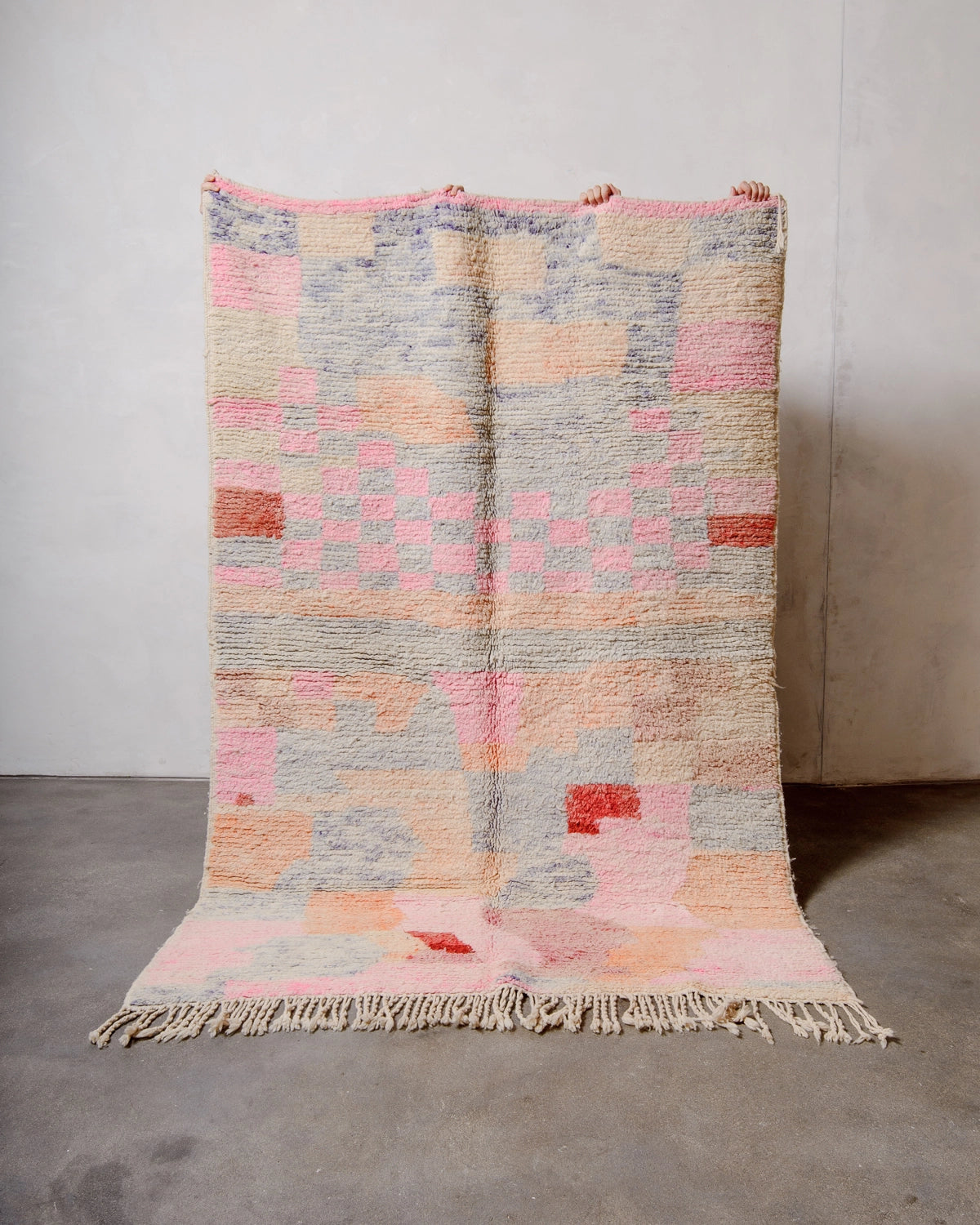 Berberlin Vintage Berber Rug made of fluffy pastel coloured wool, being held up against a white wall and grey concrete floor.